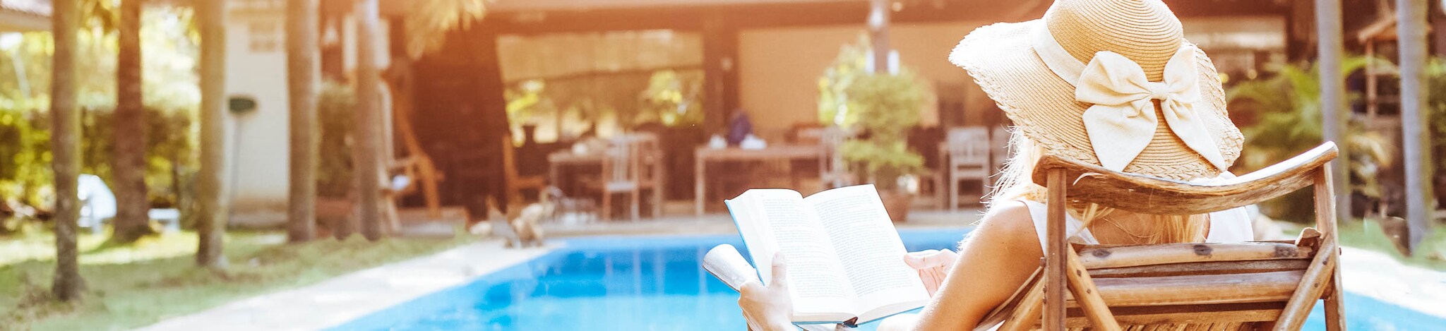 Frau liest Buch am Pool mit Ferienhaus im Hintergrund